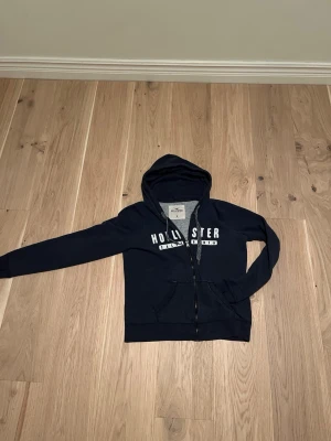 Mörkblå hoodie från Hollister - Mörkblå hoodie från Hollister med dragkedja framtill och vit text på bröstet. Klassisk huva och två fickor framtill. Tillverkad i mjukt bomullsmaterial som känns skönt mot huden. Perfekt för en avslappnad och cool stil.
