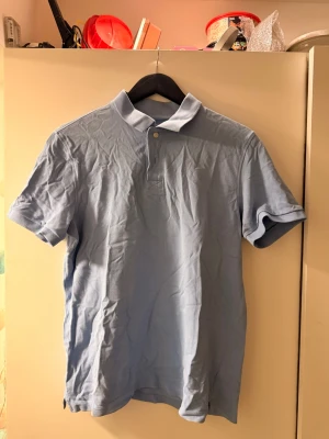 Ljusblå pikétröja i bomull - En klassisk ljusblå pikétröja med krage och två knappar framtill. Tillverkad i mjuk bomull och har korta ärmar. Perfekt för en avslappnad och stilren look.