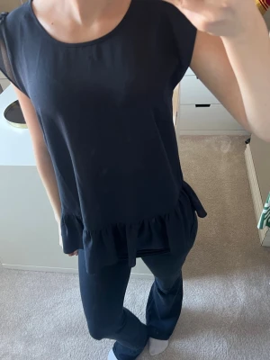 Svart volangtop med ärmlös design - Säljer en svart topp med volangkant nedtill och rund halsringning. Toppen är ärmlös och har en loose passform som ger en avslappnad vibe. Perfekt att styla med jeans eller kjol för en trendig look.