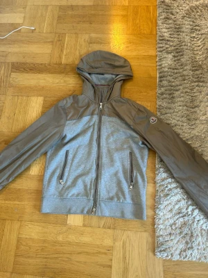 Grå vindjacka med huva från Moncler - Snygg grå vindjacka från Moncler med huva och dragkedja framtill. Jackan har kontrasterande material på ärmar och axlar, två fickor med dragkedja och Moncler-logga på ena ärmen. Perfekt för blåsiga dagar och stilren streetwear.
