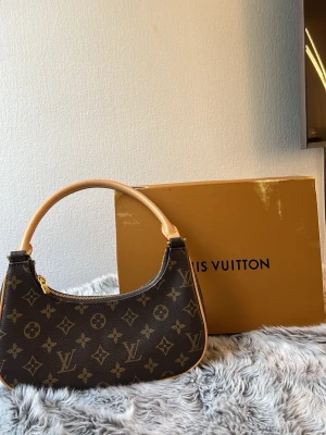  Monogram axelremsväska - Snygg handväska  i klassiskt brunt monogrammönster. Väskan har en rundad form, beige handtag och en avtagbar guldfärgad kedja. Tillverkad i canvas med läderdetaljer och dragkedja upptill. Perfekt accessoar för dig som gillar lyxig och tidlös design.