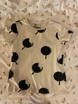 Vit t-shirt med klubbor från Lindex barn st 122/128 - Snygg vit t-shirt från Lindex med svart klubba-mönster över hela tröjan. Klassisk rund halsringning och korta ärmar. Tillverkad i mjuk bomull. Barn storlek 122/128