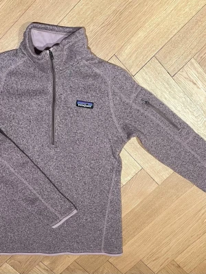 Ljuslila fleece från Patagonia - Mysig ljuslila fleece från Patagonia med halv dragkedja framtill och hög krage. Tröjan har en liten ficka med dragkedja på ärmen och klassisk logga på bröstet. Perfekt för lager-på-lager och outdoor-vibe.