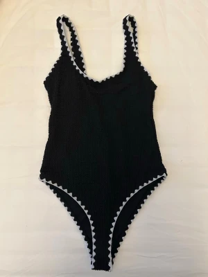 Svart baddräkt med vit kant från Boohoo - Svart baddräkt med stretchigt, smockat tyg och vita sicksack-detaljer längs kanterna från Boohoo i storlek XS. Djup rygg och klassisk skärning med breda axelband.