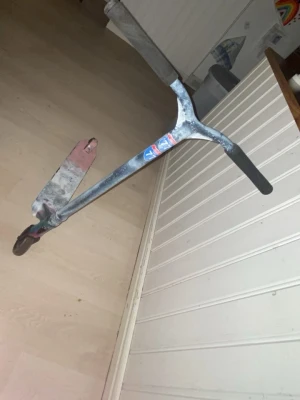 Trick sparkcykel med färgad deck - Säljer en trick sparkcykel med svart styre och handtag, samt en färgad deck i rosa, blått och grönt. Styret har klistermärken och cykeln är byggd i metall. Perfekt för skateparken eller att cruisa runt på stan.