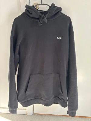 Svart hoodie från MP med logga - Säljer en svart hoodie från MP med liten vit logga på bröstet. Hoodien har en stor känguruficka framtill, ribbade muddar och en rejäl huva med dragsko. Perfekt för gym och eller avslappnad stil. Materialet känns mjukt och bekvämt.