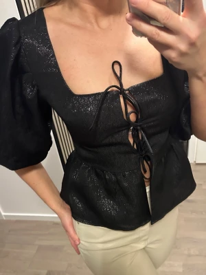 Svart glittrig topp med puffärm och snörning - Säljer en svart glittrig topp med puffärmar och snygg snörning framtill. Toppen har en fyrkantig ringning och markerad midja som ger en trendig siluett. Perfekt för dig som vill sticka ut lite extra. Materialet har en lätt glansig finish.
