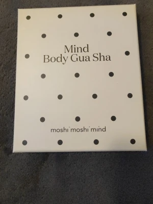 Mind Body Gua Sha från Moshi Moshi Mind - Mind Body Gua Sha från Moshi Moshi Mind är ett massageverktyg inspirerat av traditionell kinesisk teknik. Används för att stimulera blodcirkulationen och ge mjukare hud. Passar för användning på lår, rumpa, mage och armar. Kommer i en vit förpackning med svarta prickar.