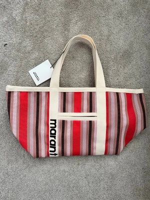 Randig shoppingväska från Marant - Snygg shoppingväska från Isabel Marant med breda handtag och färgglada ränder i rött, rosa, beige och brunt. Väskan har en praktisk ytterficka och logotypen tryckt på ena handtaget. Tillverkad i slitstarkt tygmaterial, perfekt för att bära allt du behöver.