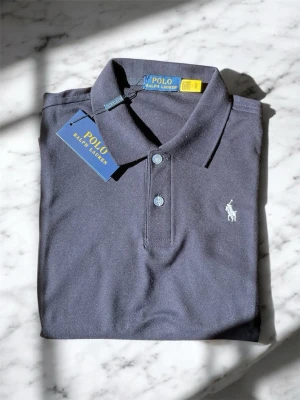 Mörkblå pikétröja från Polo Ralph Lauren - Klassisk mörkblå pikétröja från Polo Ralph Lauren med vit broderad logga på bröstet. Tröjan har krage, två knappar framtill och är tillverkad i bomull. Perfekt för en stilren och avslappnad look.