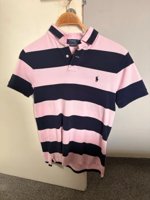 Randig pikétröja från Polo Ralph Lauren - Säljer en klassisk pikétröja från Polo Ralph Lauren i rosa och marinblått med breda ränder. Tröjan har korta ärmar, krage och knappar vid halsen samt den ikoniska broderade loggan på bröstet. Slim fit-modell i mjuk bomull. Stealerbjudande på 449 kr gäller fram till onsdag 23:59