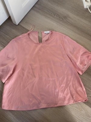 Ljusrosa blus i satin från Stockh-LM - Söt ljusrosa blus från Stockh-LM  i glansigt satinliknande material. Blusen har rund halsringning, korta ärmar och en liten öppning med knytband i nacken. Perfekt för en trendig och feminin look.