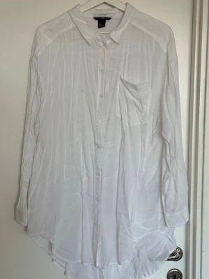 Vit långärmad skjorta från H&M - Klassisk vit skjorta från H&M i bomull med lång ärm och bröstficka. Skjortan har knappar framtill och en avslappnad passform, perfekt till många olika stilar. Enkel och tidlös design. Obs! Har en liten fläck, se bild. 