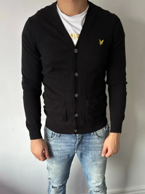 Svart cardigan från Lyle & Scott - Stilren svart cardigan från Lyle & Scott med knappstängning och två fickor framtill. Klassisk gul logga på bröstet och ribbade muddar vid ärmslut och nederkant. Perfekt att bära över en t-shirt för en avslappnad men snygg look. Använd cirka två gånger för ett år sen.