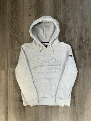 Ljusgrå hoodie från Superdry - Snygg ljusgrå hoodie från Superdry med broderad logga och japansk text på bröstet🤩 Passar storlek S-Xs och har ett litet fel som syns på sista bilden🙌🏻Mått: 62cm längd, 51cm bredd. Hör av er vid funderingar samt snabbaffär😇