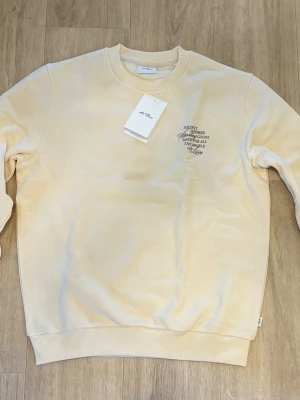 Beige sweatshirt från Les Deux - Stilren beige sweatshirt från Les Deux med tryck på bröstet och stort konstnärligt vas-motiv på ryggen. Tröjan har rund halsringning, ribbade muddar och är tillverkad i mjukt material. Perfekt för dig som gillar streetwear och snygga detaljer.