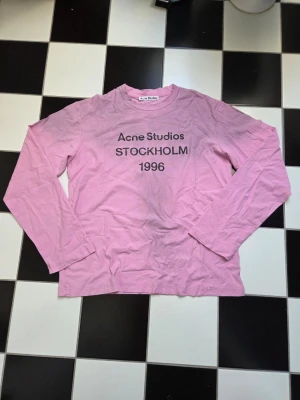 Rosa långärmad topp Acne Studios - Långärmad rosa topp från Acne Studios med tryck 'Acne Studios STOCKHOLM 1996' på bröstet. Klassisk rund halsringning och relaxed fit. Perfekt för dig som gillar minimalistisk och stilren design.