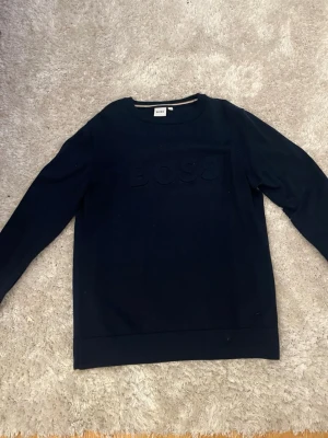Mörkblå stickad tröja från BOSS - Snygg mörkblå stickad tröja från BOSS med diskret broderad logga på bröstet. Tröjan har rund halsringning och långa ärmar.