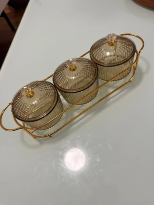 Set med tre burkar i glas och guldställ - Säljer ett set med tre runda burkar i ribbat, transparent brunt glas med lock och dekorativa gulddetaljer. Burkarna står i ett elegant guldställ med handtag, perfekt för förvaring av småsaker eller kryddor. Snygg detalj till köket eller rummet.