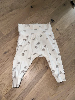 Vita byxor med igelkottsmönster - Säljer ett par vita byxor från Lindex med gulligt igelkottsmönster. Byxorna har bred mudd i midjan och vid bensluten för extra komfort. Tillverkade i mjuk ekologisk bomull, perfekta för chill dagar hemma.