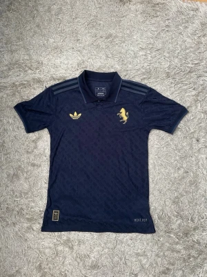 Juventus x Adidas matchtröja Vlahović - Mörkblå Juventus matchtröja från Adidas med guldiga detaljer, inklusive klubbmärke och Adidas-logga på bröstet. På ryggen står det Vlahović och nummer 9 i guld. Tröjan har pikékrage och är tillverkad i ett lätt, ventilerande material med HEAT.RDY-teknologi.