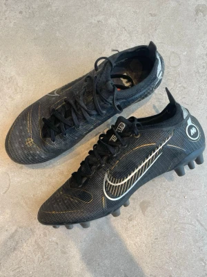 Nike Mercurial Vapor 14 svart/guld fotbollsskor Elite - Snygga Nike Mercurial Vapor 14 fotbollsskor i svart med gulddetaljer och vit swoosh. Skorna har låg profil, snörning och dobbar för spel på konstgräs (AG). Tillverkade i syntetmaterial för bra grepp och känsla på planen. Perfekta för dig som vill sticka ut på fotbollsplanen. Påse kommer med