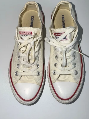 Vita Converse All Star låga sneakers - Klassiska vita Converse All Star sneakers i lågt utförande med röd och blå rand längs sulan. Skorna har canvasöverdel, vita snören och tåhätta i gummi. Perfekta för en avslappnad och tidlös stil.