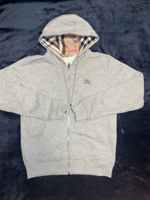 Grå zip hoodie från Burberry - Snygg grå zip hoodie från Burberry med klassiskt rutigt foder i huvan. Hoodien har dragkedja framtill, två fickor och ribbade muddar. Ikoniska Burberry-loggan broderad på bröstet. Perfekt för en avslappnad men stilren look. Storlek S men sitter nästan som en XS.
