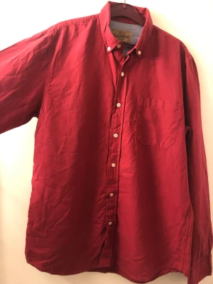 Röd skjorta med knappar - Klassisk röd skjorta med button-down krage och bröstficka. Skjortan har vita knappar framtill och långa ärmar. Tillverkad i ett mjukt bomullstyg som känns skönt mot huden. Perfekt för dig som gillar en enkel men snygg stil.