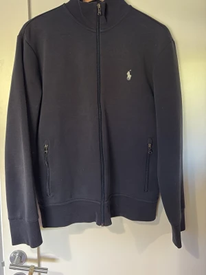 Marinblå ziptröja från Ralph Lauren - Snygg marinblå ziptröja från Ralph Lauren med hög krage och dragkedja framtill. Tröjan har två fickor med dragkedja och den klassiska vita logon broderad på bröstet. Tröjan är i väldigt bra skick inget som är sönder på den. Den är lätt använd då jag inte riktigt trivdes i den. Pris går att diskutera, hör av er vid minsta funderingar😊