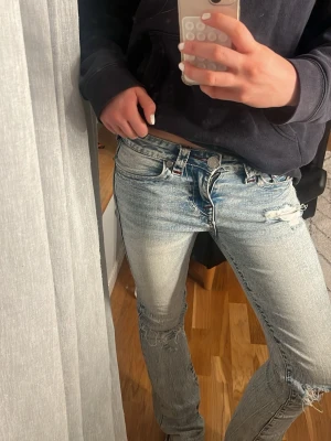 Ljusblå jeans True Religion - Säljer ett par ljusblå jeans från True Religion med slitningar och hål på benen för en trendig look. Jeansen har klassisk femficksmodell, låg midja och kontrastsömmar på bakfickan. Perfekta för dig som gillar streetstyle och avslappnad vibe.