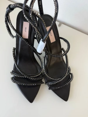 Svarta klackar med strass från Nelly - Säljer ett par svarta pumps från Nelly med spetsig tå och tunna remmar dekorerade med svarta strasstenar. Perfekta för att lyfta din outfit med lite extra bling. 