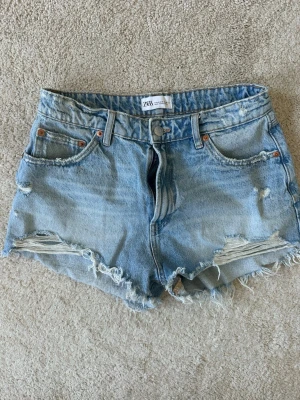 Blå jeans shorts från Zara - Säljer ett par ljusblå jeansshorts från Zara, fransiga kanter och slitna detaljer framtill. Klassisk femficksmodell med knappgylf. Perfekta för sommardagar och har en avslappnad, loose passform.