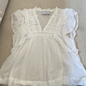 Vit volangblus  -  vit blus från neo noir med v-ringning, volangdetaljer. Luftig och lätt i materialet, perfekt för varma dagar. Ärmlös design med breda volangärmar ger en feminin och bohemisk känsla.