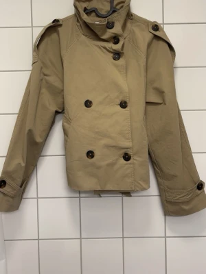 Kort beige trenchcoat med knytband - ANDVÄND BARA 1 GÅNG!!!     (Buda gärna)                             Stilren beige trenchcoat i kort modell med dubbelknäppning, axelklaffar och knytband i midjan. Jackan har stora knappar framtill och vid ärmsluten samt en hög krage. Tillverkad i ett slitstarkt bomullsliknande material.