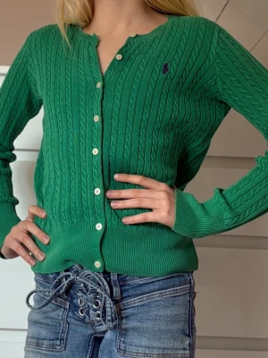 Grön kabelstickad kofta från Ralph Lauren - Grön kabelstickad kofta från Ralph Lauren med lila broderad logga på bröstet. Koftan har långa ärmar, ribbade muddar och knäppning med vita knappar framtill. Perfekt för lager på lager och en klassisk preppy stil.