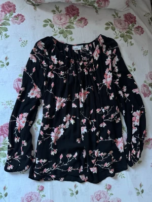 Svart blommig blus - Svart blus med rosa och vita blommor. Blusen har lång ärm, rundad halsringning och ett luftigt, lätt tyg som ger en avslappnad känsla. Perfekt för dig som gillar romantiska mönster och vill ha något bekvämt men ändå snyggt.