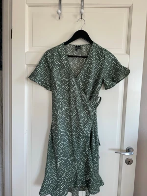 Grön omlottklänning med prickigt mönster - Grön omlottklänning med vitt prickigt mönster från veromoda. Klänningen har korta ärmar, knyt i midjan och v-ringad hals. Har använts några fåtal gånger men har inga defekter. Storlek xs💕