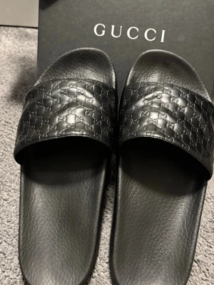 Svarta slides från Gucci - Säljer ett par svarta slides från Gucci med klassiskt GG-mönster på remmen. Tillverkade i slitstark gummi med strukturerad sula och bred rem över foten. Perfekta för en avslappnad och trendig look.