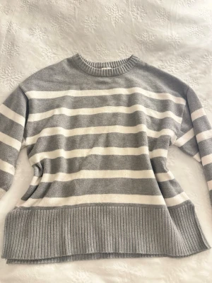 Gråvit randig stickad tröja - Säljer en stickad tröja med breda horisontella ränder i grått och vitt. Tröjan har rund halsringning, långa ärmar och bred ribbstickad kant nedtill. Perfekt för dig som gillar en avslappnad och stilren look.
