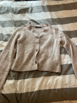 Beige kofta med knappar - Mysig beige kofta med v-ringning och tre knappar framtill. Koftan har lång ärm och är tillverkad i ett mjukt, stickat material. Perfekt att slänga över en t-shirt eller linne för en avslappnad look.