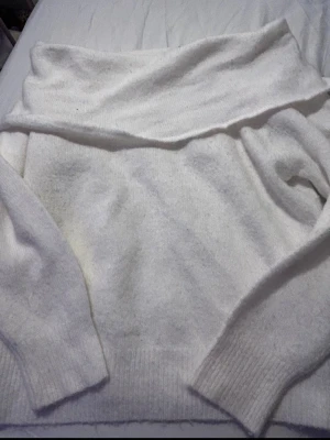 Vit off shoulder stickad tröja - Säljer en vit stickad tröja med bred off shoulder-krage och långa ärmar. Tröjan har en mjuk och fluffig känsla, perfekt för att hålla värmen. Ribbstickade muddar vid ärmslut och nederkant ger en snygg finish.