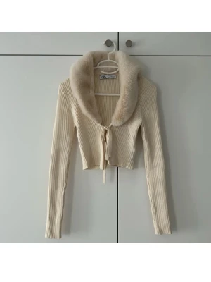 Beige ribbad kofta med fuskpäls från Zara - Trendig beige ribbad kofta från Zara med croppad passform och långa ärmar. Koftan har en bred, mjuk fuskpäls-krage och knyts med ett band framtill. Perfekt för dig som vill ha en statement piece i garderoben. Den är ny utan prislapp