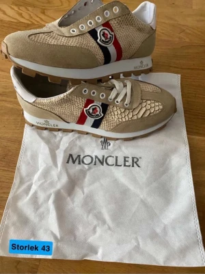 Beige sneakers från Moncler, strl 43 - Snygga beige sneakers från Moncler med mockadetaljer och texturerad ovandel i reptilmönster. Skorna har vita snören, Moncler-logga på sidan och röd/blå/vit rand. Yttersulan är grovt mönstrad för extra grepp. Kommer med original Moncler dustbag.