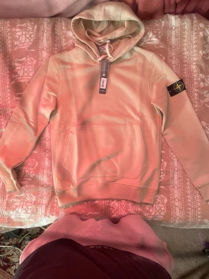 Beige hoodie från Stone Island - Snygg beige hoodie från Stone Island med klassisk svart och gul logotyp-patch på vänster ärm. Hoodien har huva med dragsko och en stor magficka framtill. Tillverkad i mjukt material som känns skönt mot huden. Perfekt för en chill och stilren look.