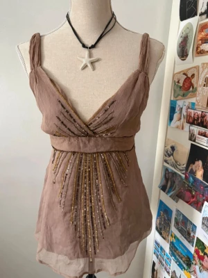 Beige linnetopp med pärlor - Snygg beige linnetopp med breda axelband och v-ringning. Toppen har dekorativa pärlor och broderade detaljer framtill i guld och brunt. Lätt transparent material och knäppning med knappar bak. Perfekt för dig som gillar unika detaljer.