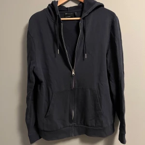 Svart hoodie med dragkedja Asos design - Svart hoodie från Asos design med hel dragkedja framtill, huva med snörning och två stora fickor. Tröjan är långärmad och har en loose passform, perfekt för chill dagar. Materialet är mjuk bomull som känns skönt mot huden.