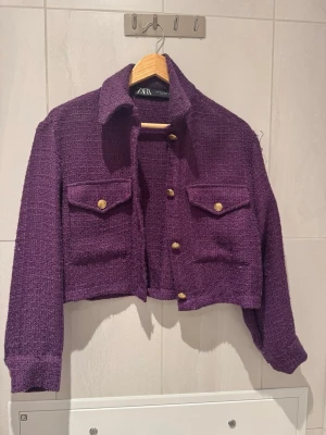 Lila kvaj från Zara - Trendig lila kavaj  från Zara i grovstickat material. Kort modell med krage, två bröstfickor med guldknappar och långa ärmar. Perfekt för att lyfta din outfit med en färgklick och cool retrovibe.