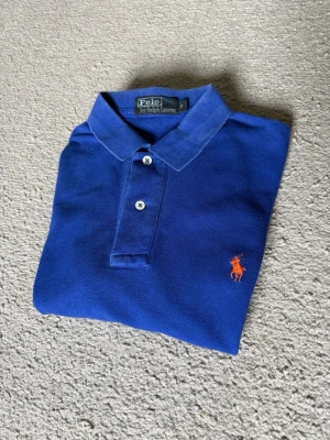 Blå pikétröja från Polo Ralph Lauren - Klassisk blå pikétröja från Polo Ralph Lauren med orange broderad logga på bröstet. Tröjan har korta ärmar, krage och två knappar framtill. Tillverkad i bomull för en bekväm känsla och snygg passform.
