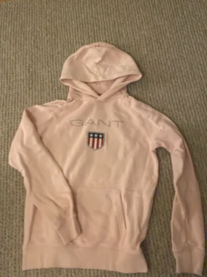 Ljusrosa hoodie från GANT - Säljer en ljusrosa hoodie från GANT med broderad logga och sköld på bröstet. Hoodien har huva och en stor magficka framtill. Perfekt för en avslappnad och stilren look. Materialet känns mjukt och bekvämt.